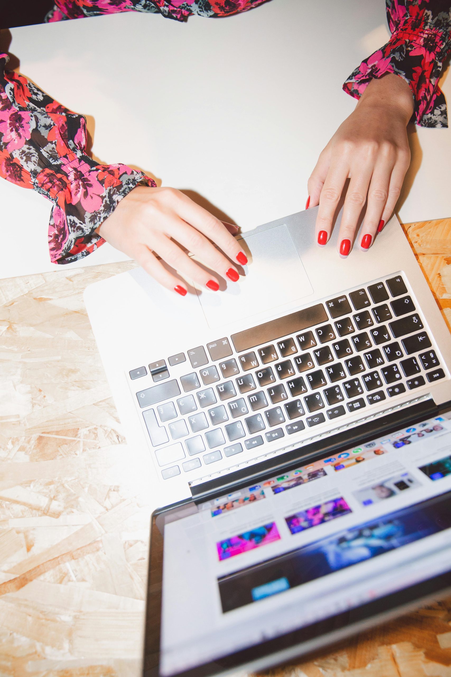 Overhead view of woman using laptop, showcasing red manicured nails and floral blouse in a modern workspace.